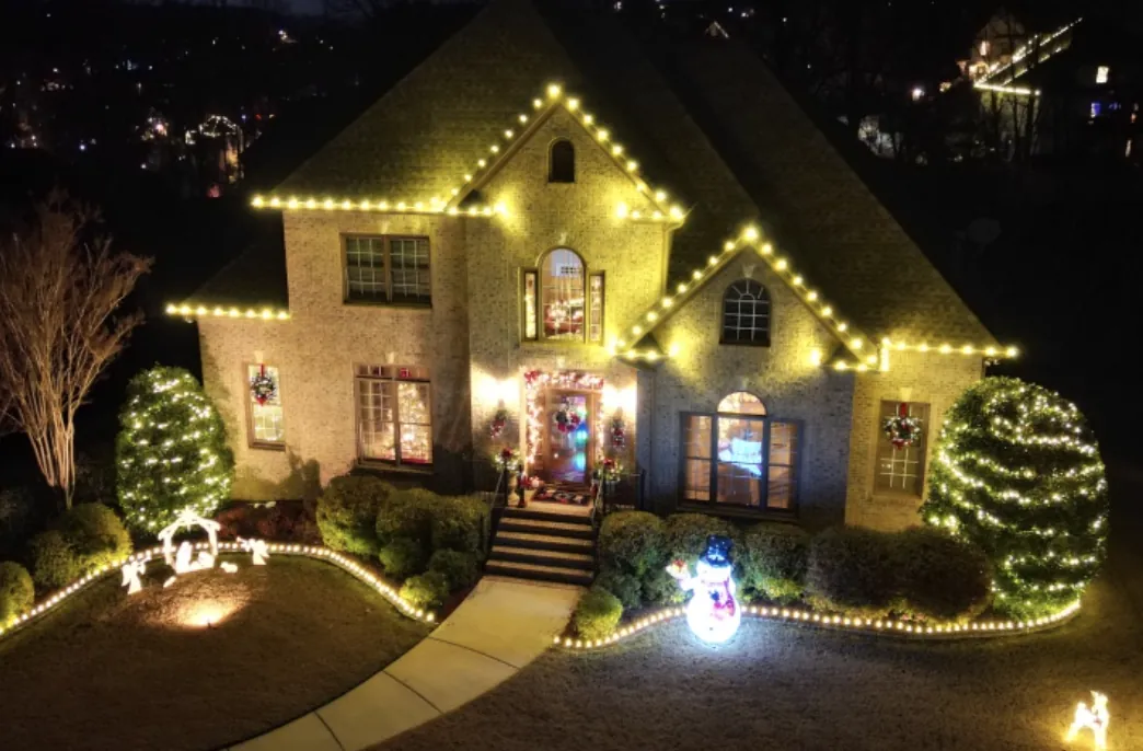 Elevated view of a brick home with roofline lights, wreaths, wrapped shrubs and lit walkway edging.