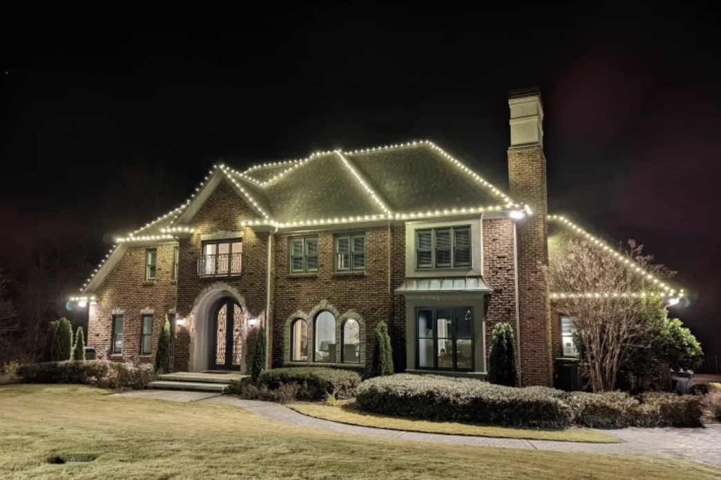 Roofline lights on a large brick home with a chimney and shaped front shrubs at night.