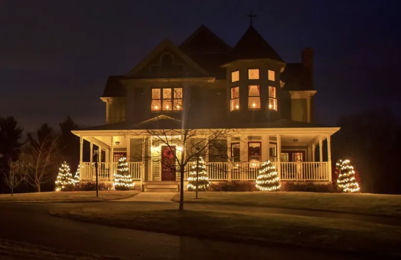 Small trees wrapped in white lights in front of a Victorian-style home with a wraparound porch.